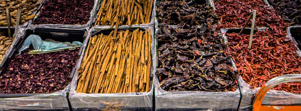 Colorful bins of dried spices at a market, including cinnamon sticks, dried chilies, and hibiscus petals.
