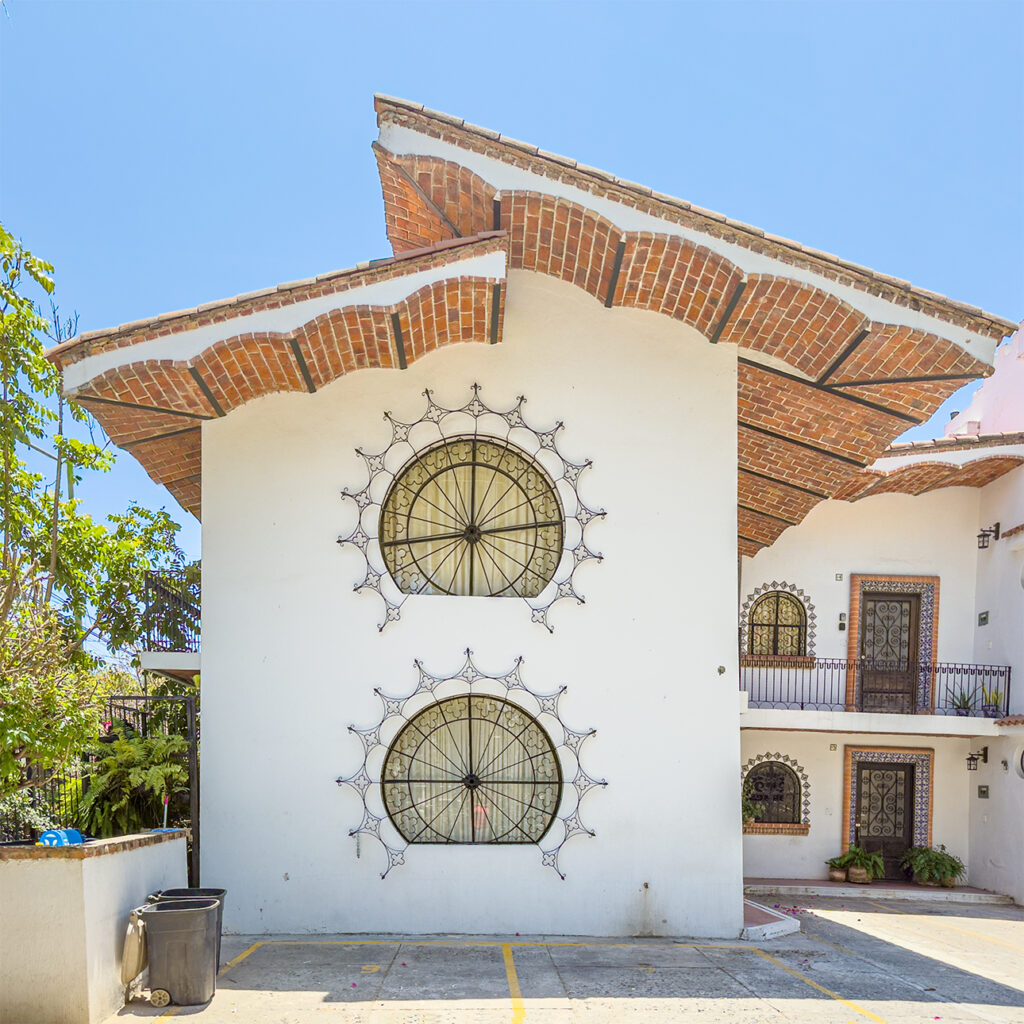 White adobe building with round windows framed in decorative ironwork, and a dramatic tiled roof against a clear blue sky.