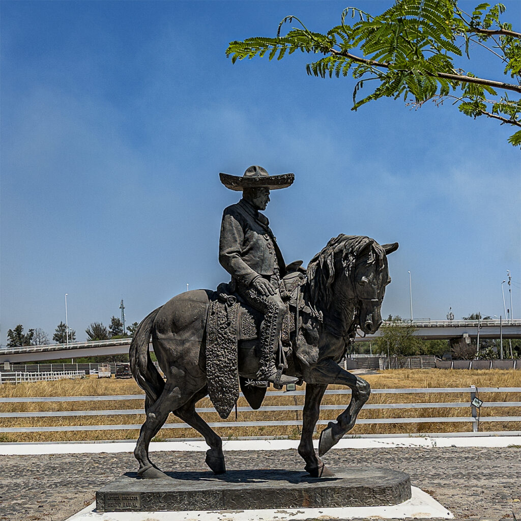 Statue of a charro in traditional dress and sombrero, riding a horse with intricate tack, set against a dry landscape.