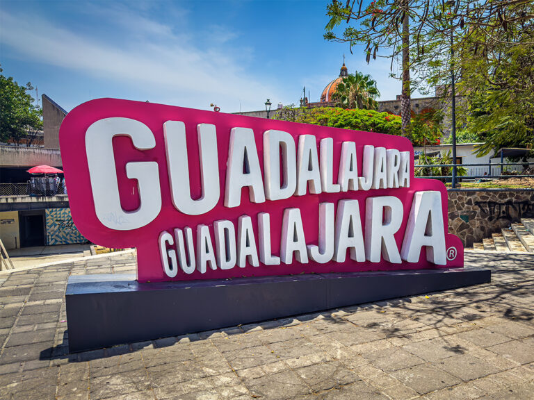 Large pink and white “GUADALAJARA GUADALAJARA” sign in a public square with trees with a cathedral dome in the background.