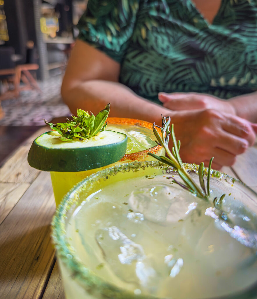 Two mezcal cocktails on a rustic table, Tammi’s hands in the background, waiting to grab hers.