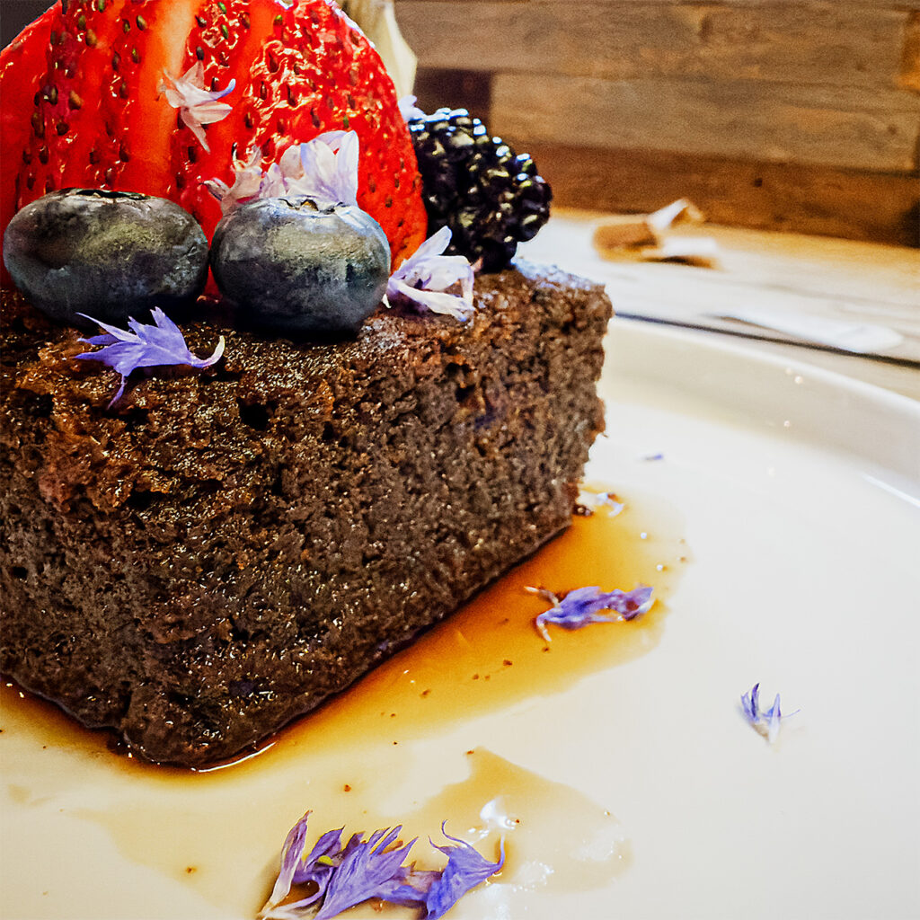 Chocolate cake topped with fresh berries and purple edible flowers, soaked in Mezcal syrup