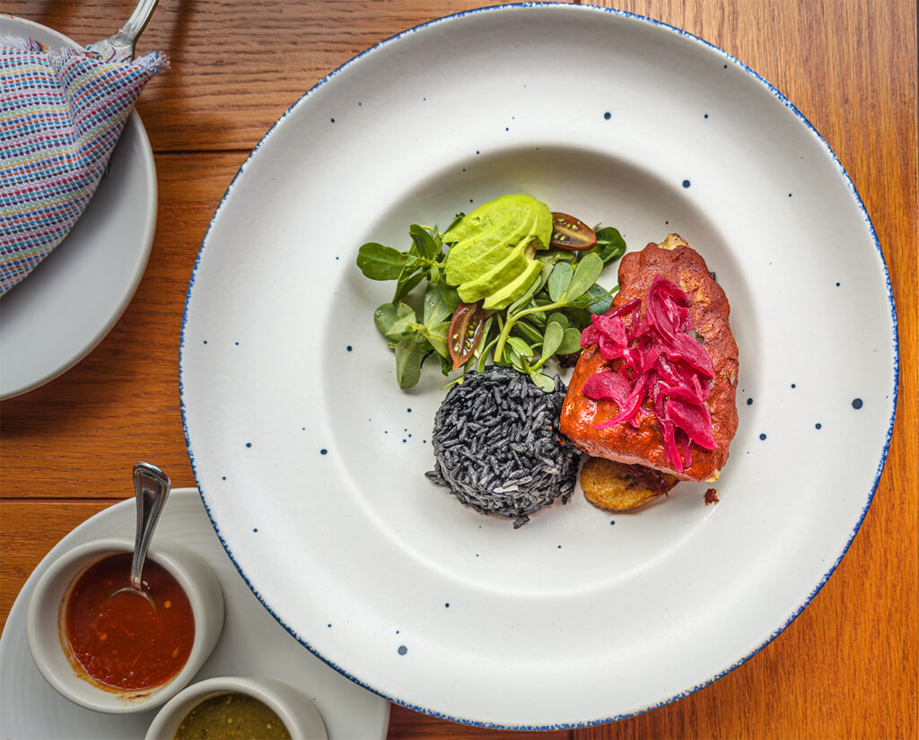 Elegant dish featuring black rice, avocado atop fresh greens, and fish topped with vibrant pickled onions