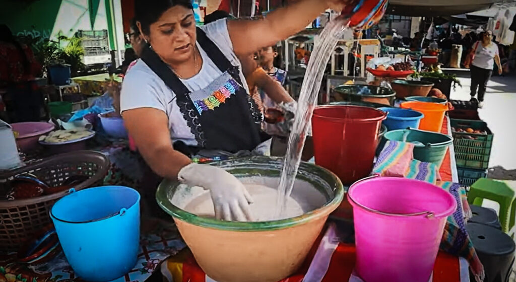 A woman in a traditional apron hand-mixing tejate in a large clay bowl at a bustling Oaxacan market,